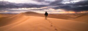 Un homme marche seul sur une dune de sable devant un ciel chargé de nuages noirs